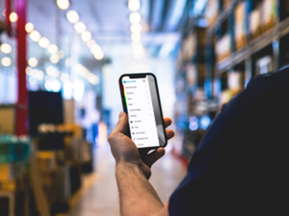 A person in a warehouse holding a cellphone displaying Monitor Mobile menu. 