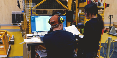Two people standing in a manufacturing environment looking at a computer displaying a Monitor ERP dashboard.