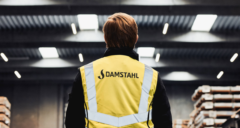 A person wearing a yellow vest with the text “Damstahl” stands inside a warehouse.