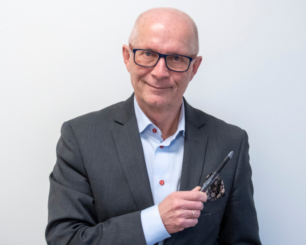 Portrait of an older man in a gray suit, wearing glasses and holding a pen in his right hand. He has a light smile and a patterned pocket square in his jacket, standing against a plain white background.