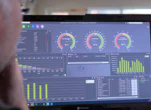 A person analyzing data displayed on a computer monitor showing various graphs, charts, and gauges. The screen includes bar graphs, line graphs, and circular gauges, indicating performance metrics or system monitoring data.