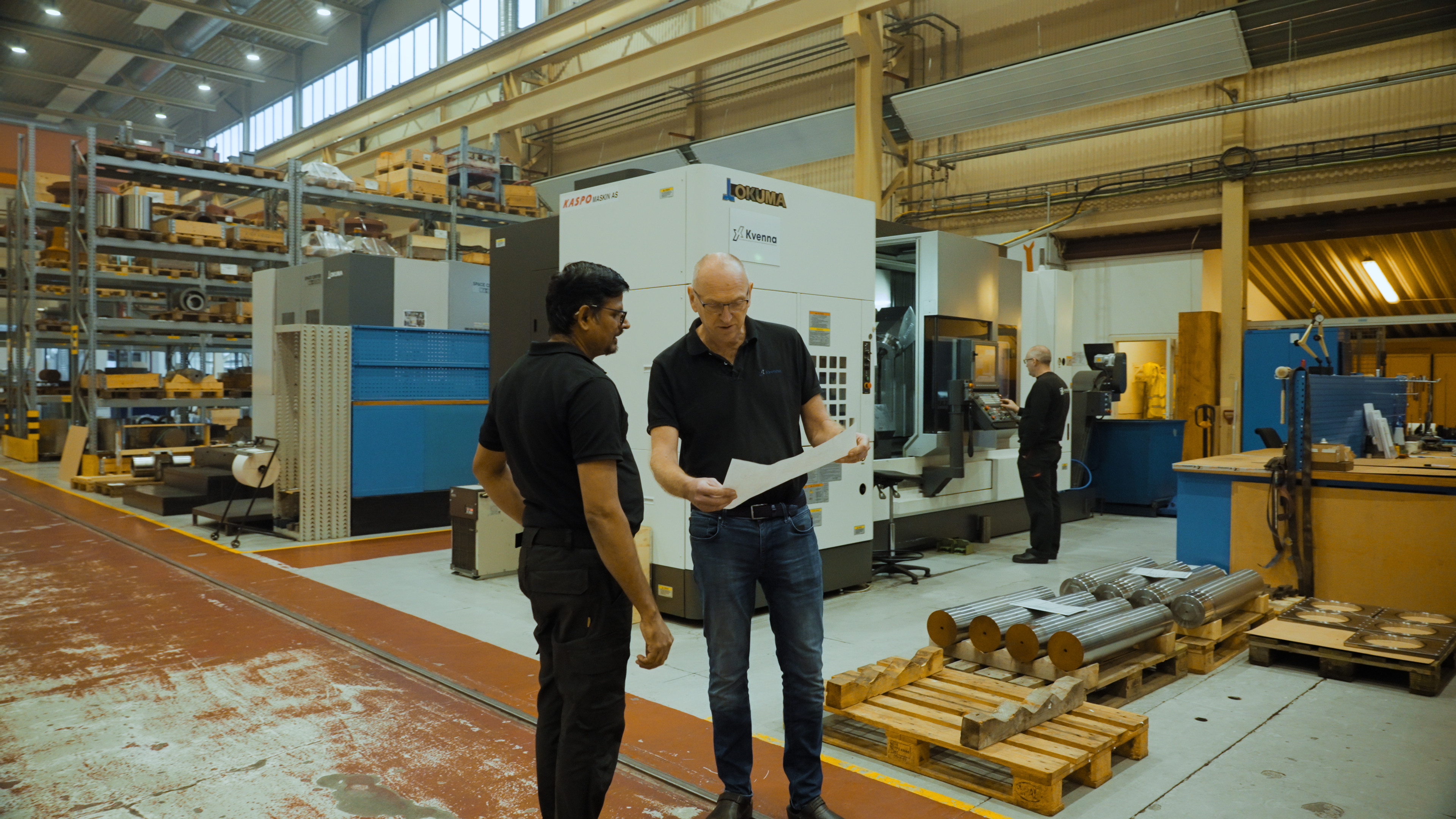 To menn i svart arbeidstøy står i en industrihall og ser på et dokument. I bakgrunnen er en stor maskin med teksten ‘Okuma’, samt hyller fylt med utstyr og en medarbeider som jobber ved en maskin. Flere metallstenger ligger stablet på paller i forgrunnen.