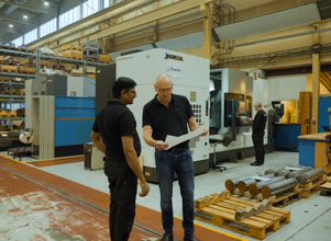 To menn i svart arbeidstøy står i en industrihall og ser på et dokument. I bakgrunnen er en stor maskin med teksten ‘Okuma’, samt hyller fylt med utstyr og en medarbeider som jobber ved en maskin. Flere metallstenger ligger stablet på paller i forgrunnen.