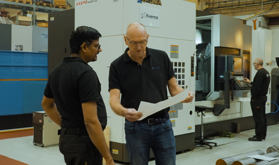 To menn i svart arbeidstøy står i en industrihall og ser på et dokument. I bakgrunnen er en stor maskin med teksten ‘Okuma’, samt hyller fylt med utstyr og en medarbeider som jobber ved en maskin. Flere metallstenger ligger stablet på paller i forgrunnen.