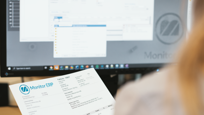 A person holding an invoice  in front of a computer screen while checking information in Monitor ERP.