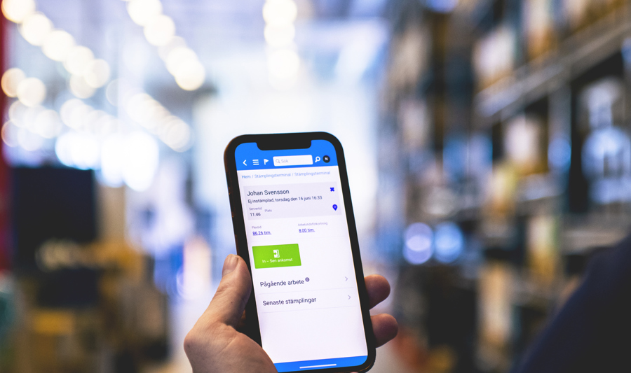 The image shows a person holding a smartphone with the screen visible, displaying an app interface, in a blurred retail or warehouse setting, suggesting the use of mobile technology for shopping or inventory management.